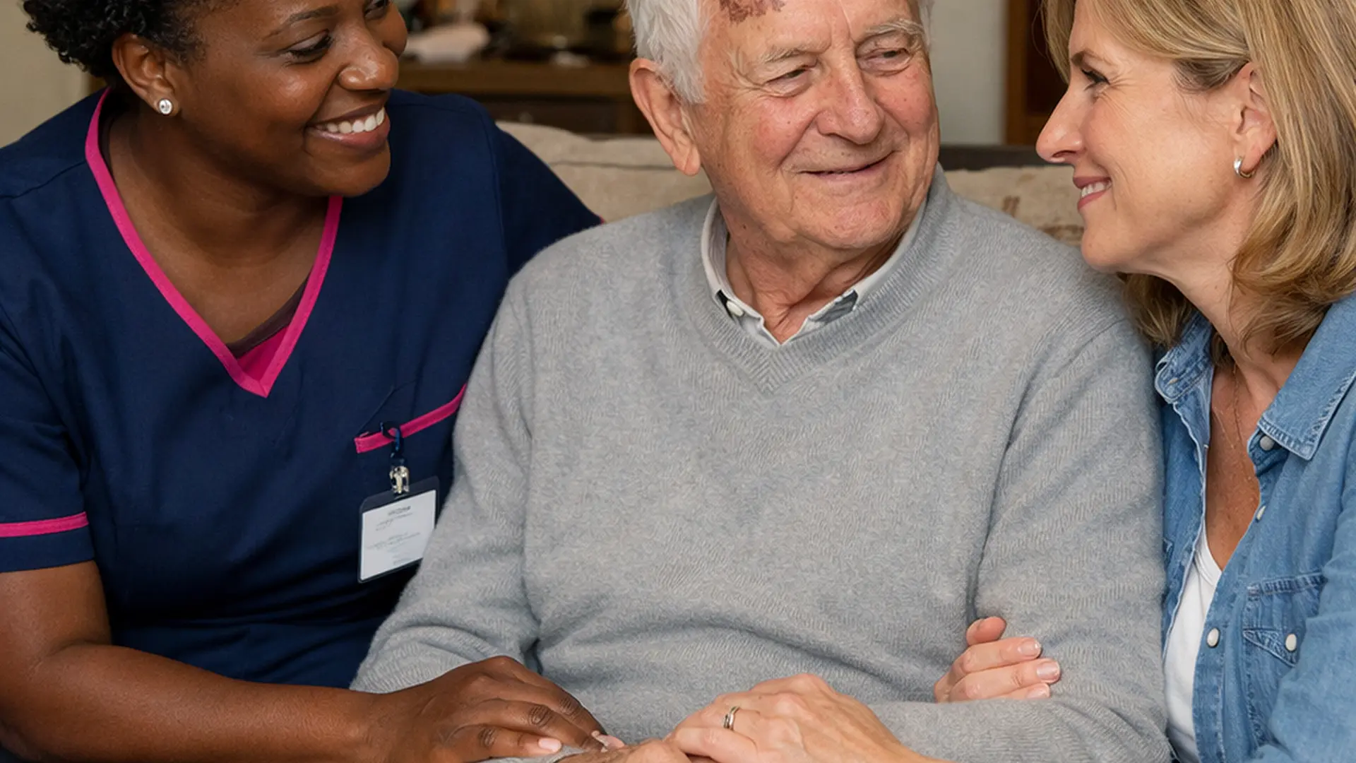 A realistic and tender home care scene of a black female caregiver in a 00072b tunic with e51974 accents sitting beside an elderly caucasian man and his daughter sharing a reassuring conversation in a cosy lounge all appearing understood calmer and supported after a fall.