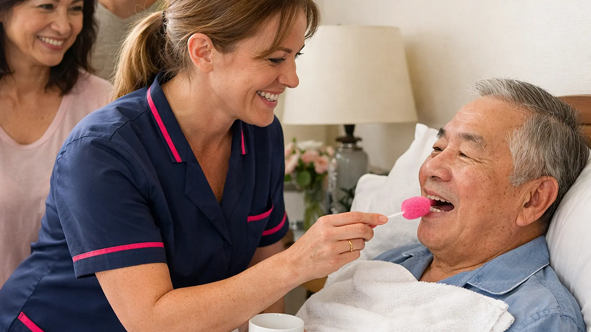 A realistic care at home image with a caucasian female caregiver in a tunic uniform colour 00072b with e51974 accent gently providing mouth care and freshening bedding for an asian elderly male service user warm smiles from relatives bright but soft room respectful and compassionate atmosphere
