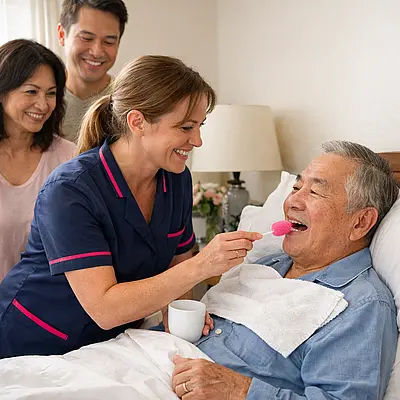 A realistic care at home image with a caucasian female caregiver in a tunic uniform colour 00072b with e51974 accent gently providing mouth care and freshening bedding for an asian elderly male service user warm smiles from relatives bright but soft room respectful and compassionate atmosphere