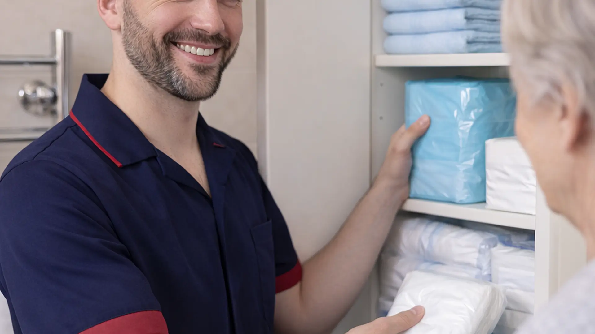 A realistic close up view in a tidy ensuite bathroom where a caucasian male caregiver in a 00072b tunic with e51974 accents arranges discreet continence products in a cabinet while chatting reassuringly with an unseen older person creating a sense of organisation privacy and calm support.