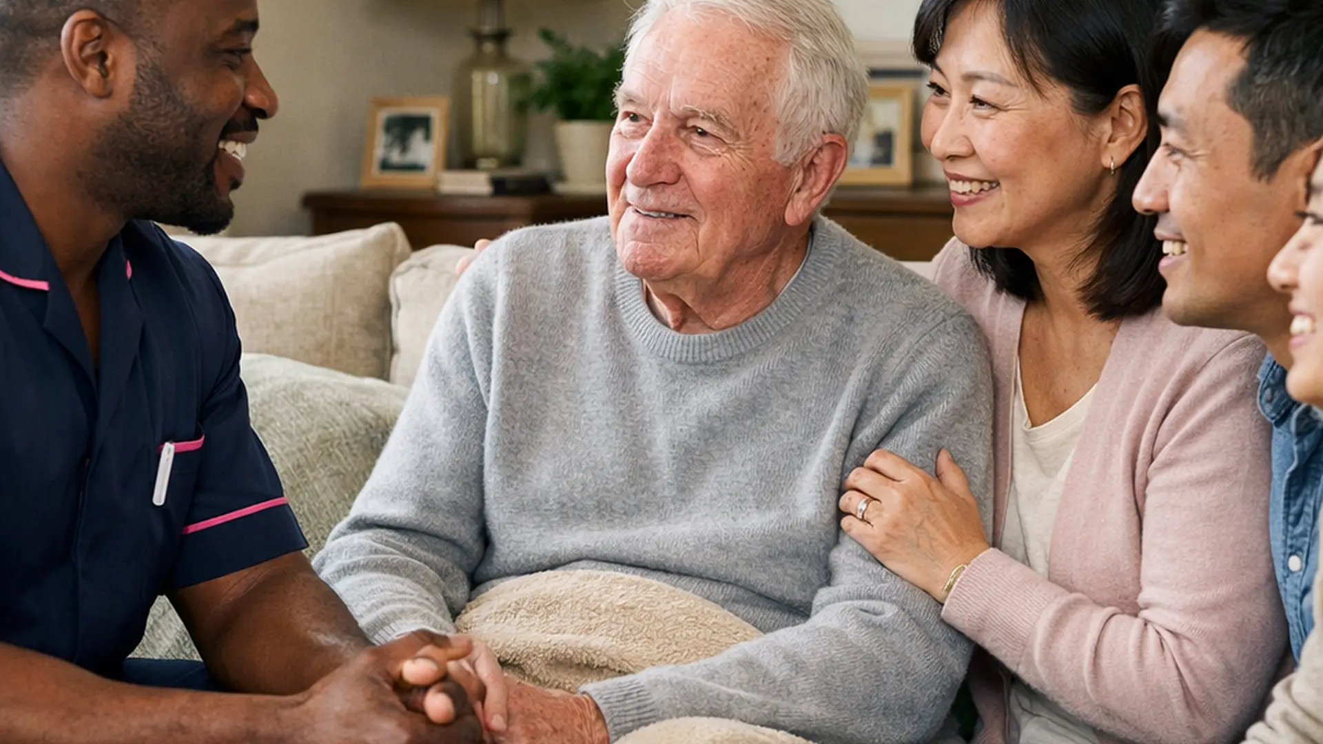 A realistic front room home care scene with a black male caregiver in a 00072b tunic with e51974 accent holding a reassuring conversation with an asian family and an elderly caucasian service user all smiling softly with natural lighting comforting dcor and a strong sense of trust warmth and dignity