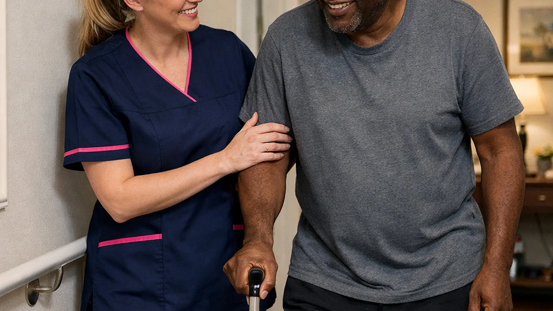 A realistic scene of a caucasian female caregiver in a 00072b tunic with e51974 accent helping a black male service user walk carefully through his hallway after surgery. Both appear relaxed and encouraged with handrails and a clean safe home environment conveying reassurance and recovery at home.