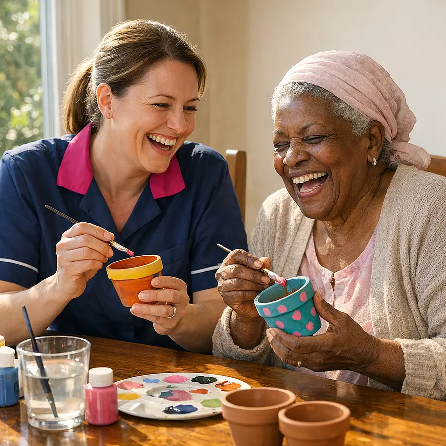 Caucasian female caregiver in a 00072b tunic with e51974 collar sitting at a dining table with an older Black woman both laughing while painting small plant pots sunlight streaming through the window emphasising joy in hobbies despite illness.