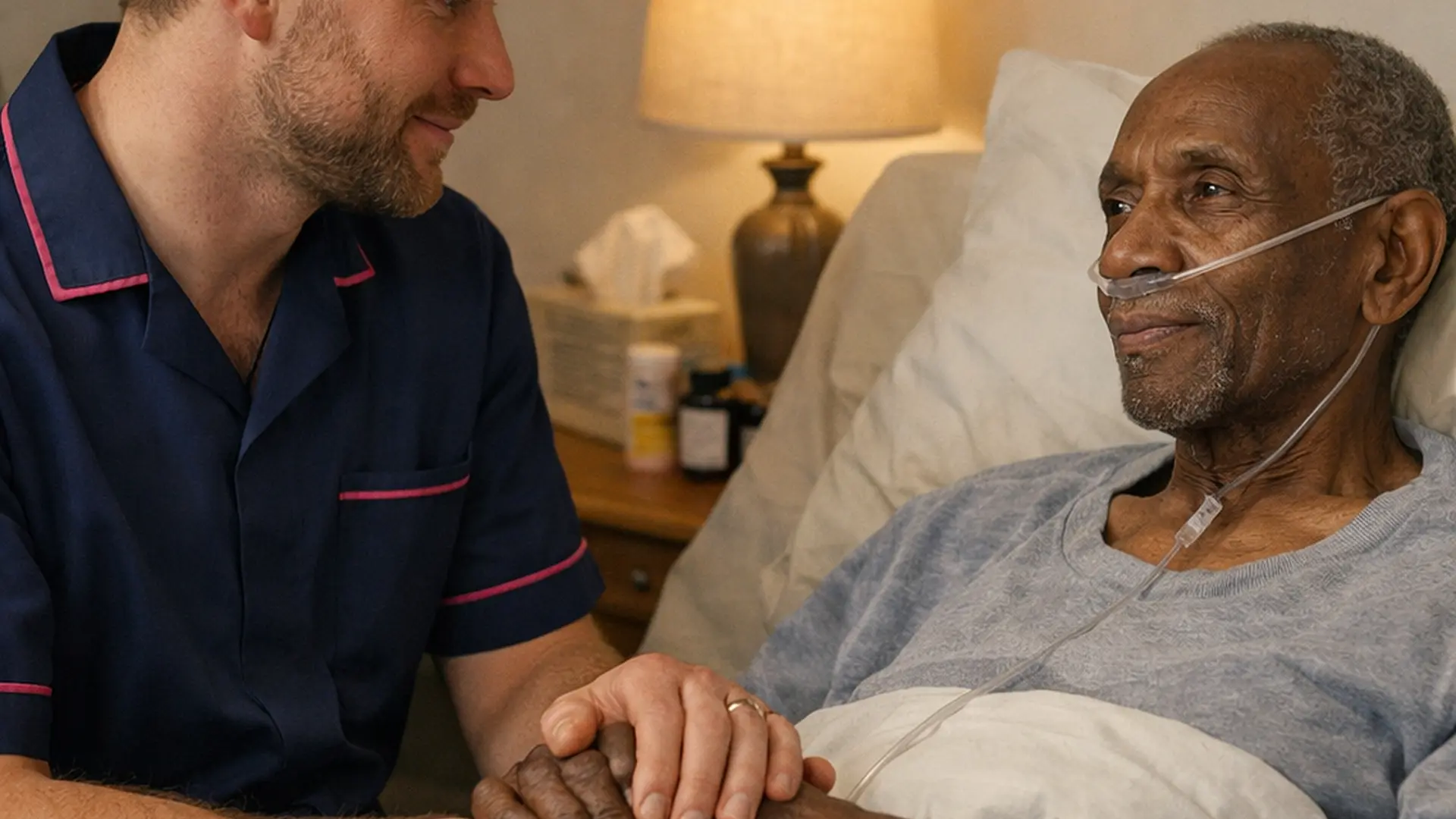Gentle realistic scene of a white male caregiver in a 00072b tunic with e51974 piping sitting at the bedside of a frail black man with oxygen tubing holding his hand in a softly lit bedroom emphasising advanced lung cancer support.