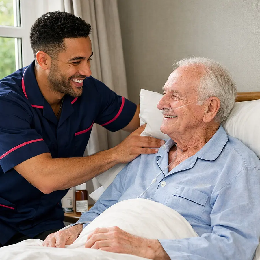 Realistic image of a mixed race male caregiver in a 00072b tunic with e51974 accents adjusting pillows behind a frail white older man sitting up in bed by a window both sharing a reassuring smile showing lung cancer care being delivered at home.