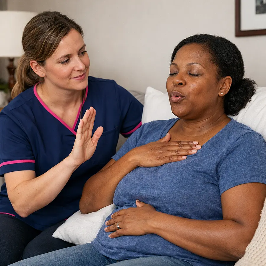 Realistic image of a white female caregiver in a 00072b tunic with e51974 accents helping a tired black woman sit upright with cushions on a sofa demonstrating breathing techniques with calm expressions.