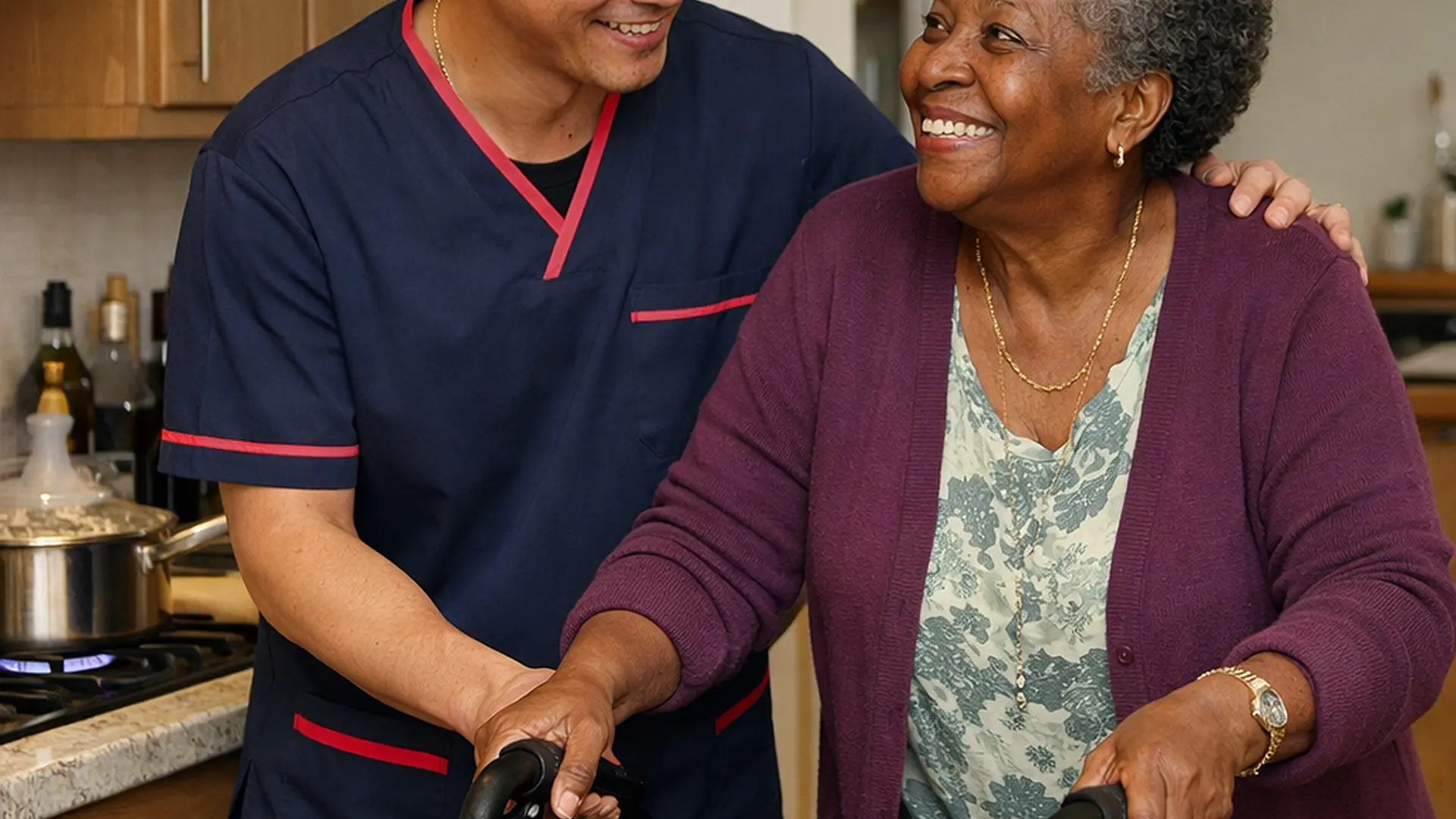 Realistic image of an Asian male caregiver in a 00072b tunic with e51974 accents supporting an older black woman as she uses a rollator in her kitchen a pot simmering on the hob both smiling demonstrating integrated mobility and daily living support.