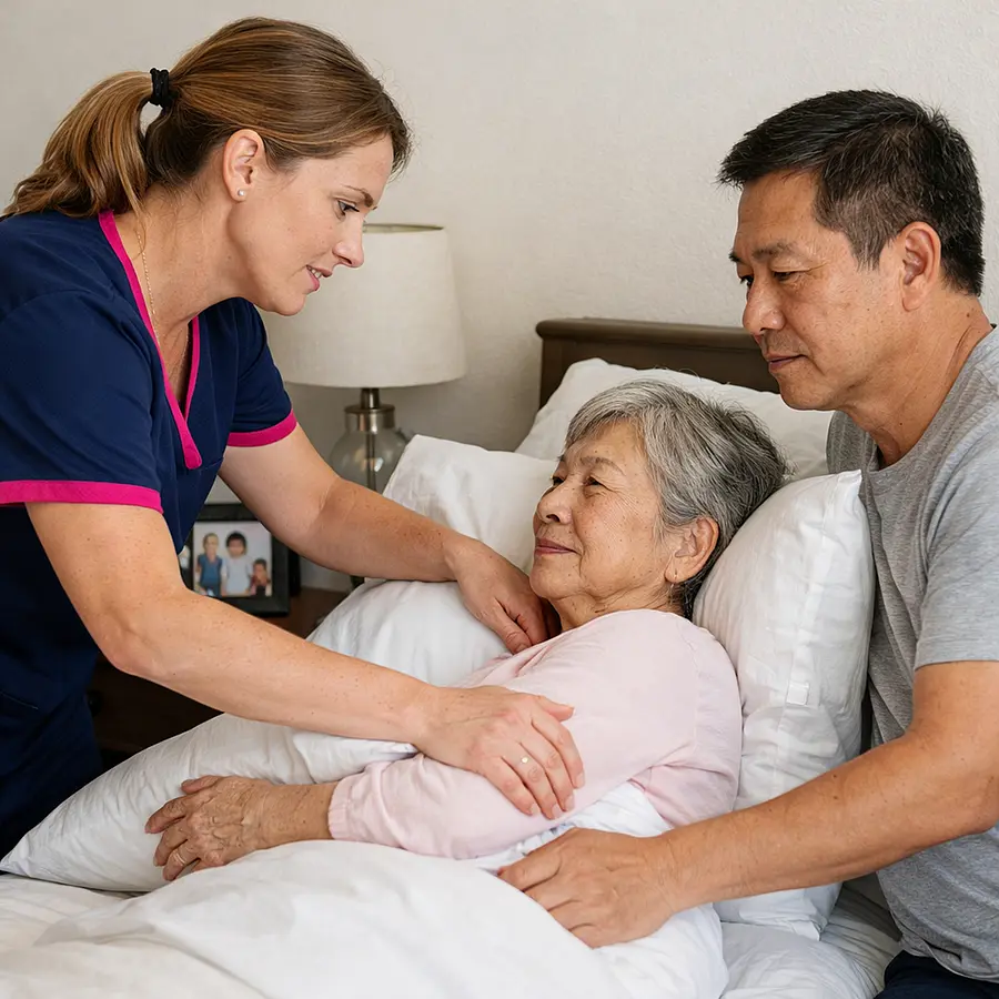 Realistic scene of a white female caregiver in a 00072b tunic with e51974 trim demonstrating to a middle aged Asian man how to gently reposition his elderly mother in bed using pillows all three focused but relaxed clean bedding and a nightstand with family photos nearby.