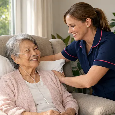 Realistic scene of a white female caregiver in a 00072b tunic with e51974 piping gently adjusting cushions for an elderly Asian woman seated in a reclining chair oxygen tubing visible sun streaming through window houseplants nearby both sharing a soft smile showing comfort and understanding.
