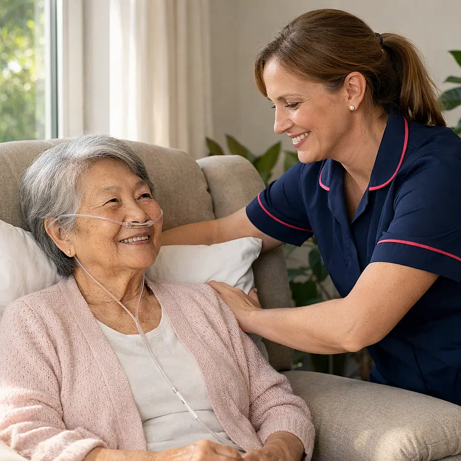 Realistic scene of a white female caregiver in a 00072b tunic with e51974 piping gently adjusting cushions for an elderly Asian woman seated in a reclining chair oxygen tubing visible sun streaming through window houseplants nearby both sharing a soft smile showing comfort and understanding.