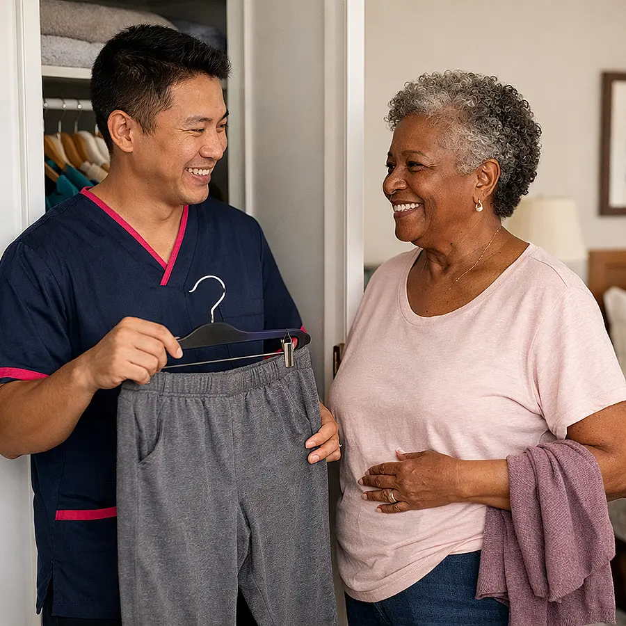 Warm realistic photo of an Asian male caregiver in a 00072b tunic with e51974 trim helping an older Black woman choose clothing from her wardrobe considering comfort around her stoma both smiling and talking bedroom tidy and bright.