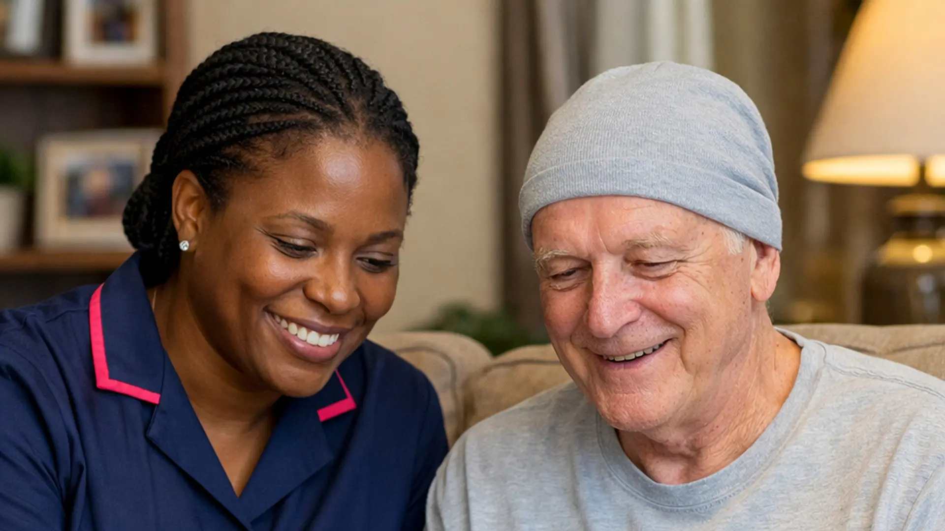 Warm, realistic scene of a Black female caregiver in a navy #00072b tunic with #e51974 accents, sitting beside a white male client in his living room, both smiling as they look at some paperwork about his cancer treatment, conveying reassurance and calm home support.
