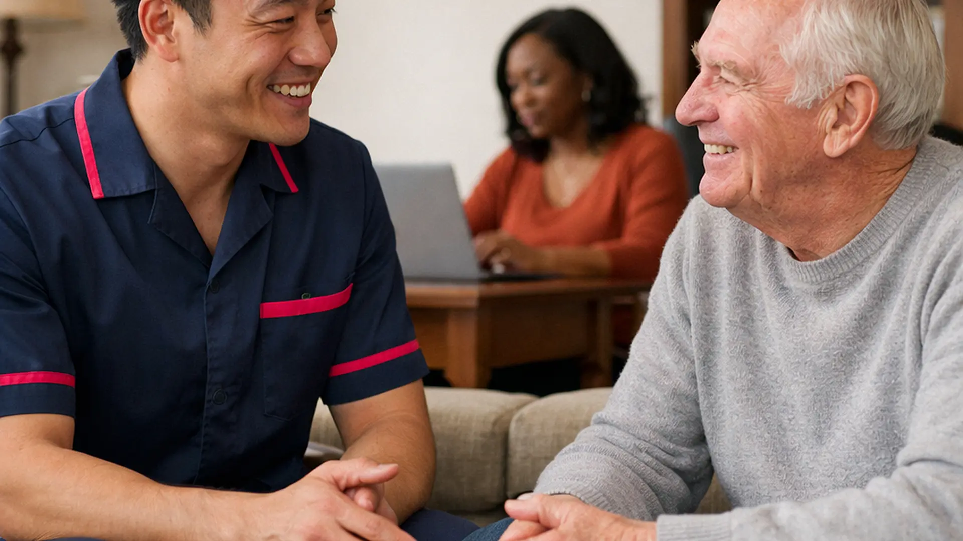 realistic asian male caregiver in tunic uniform 00072b with e51974 accent chatting warmly with a caucasian male service user while a black female family carer works calmly on a laptop in the same home professional and homely joyful reassurance realistic photo style